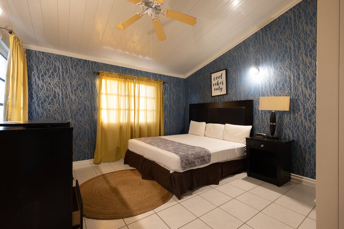 Une chambre à coucher avec un lit, une table de chevet, un ventilateur de plafond et une fenêtre avec des rideaux.