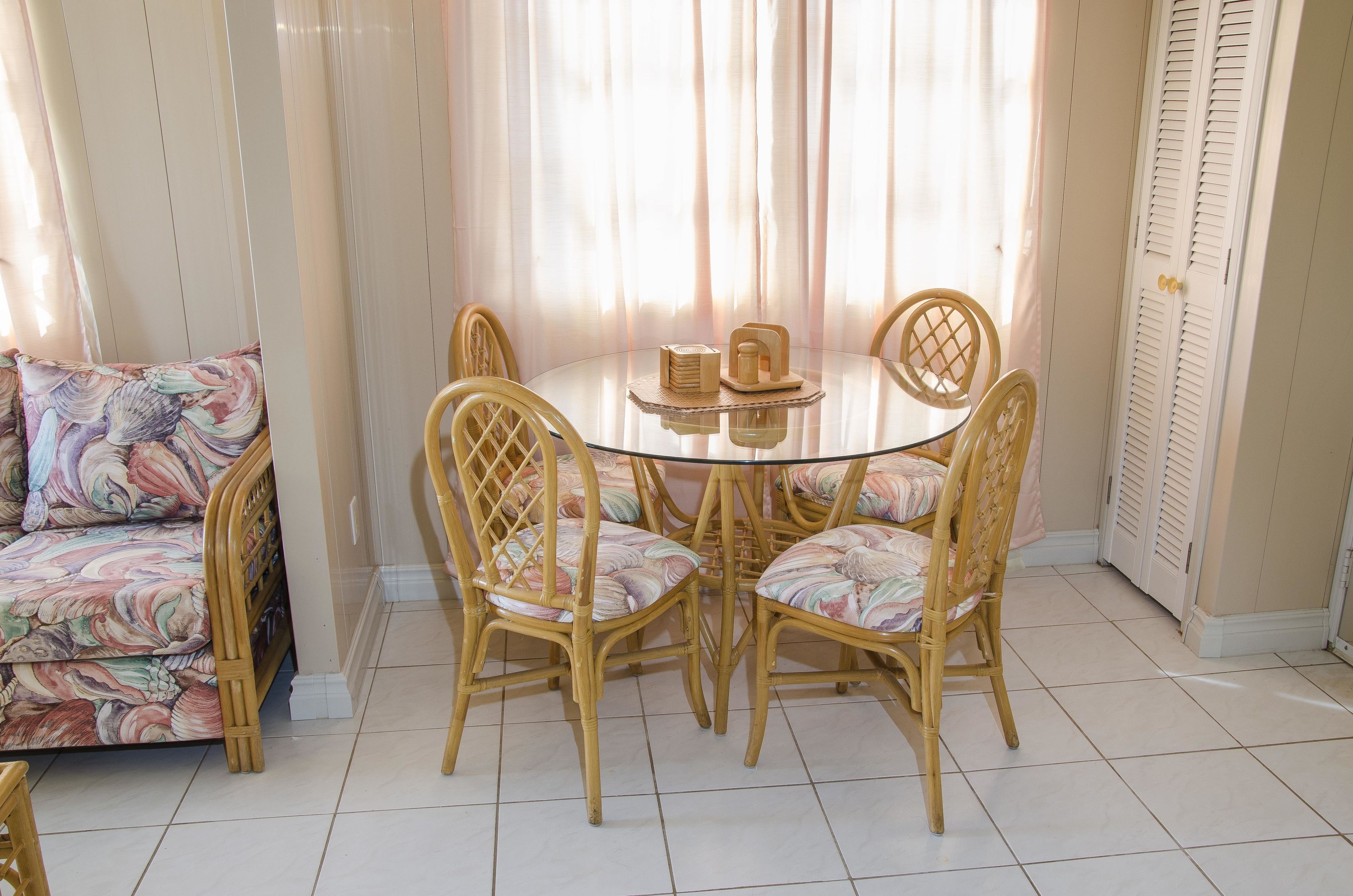 Uma área de refeições com uma mesa de vidro redonda e quatro cadeiras de vime, um sofá com uma almofada colorida e uma janela com cortinas leves.