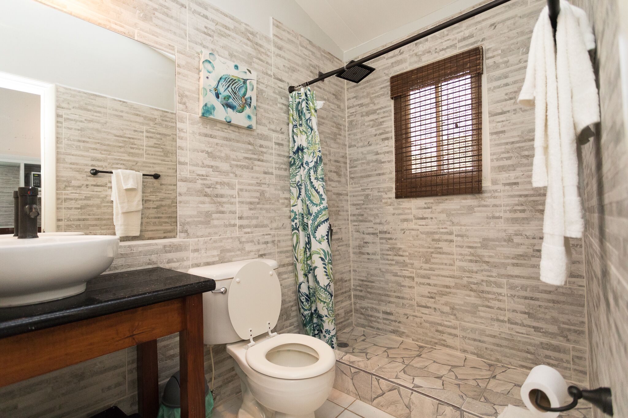 Un baño con inodoro blanco, lavamanos con encimera negra, ducha con cortina estampada y ventana con persianas.