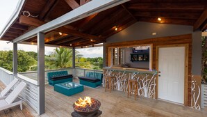 Un espacio al aire libre techado con suelo de madera, una chimenea, asientos y una zona de bar.