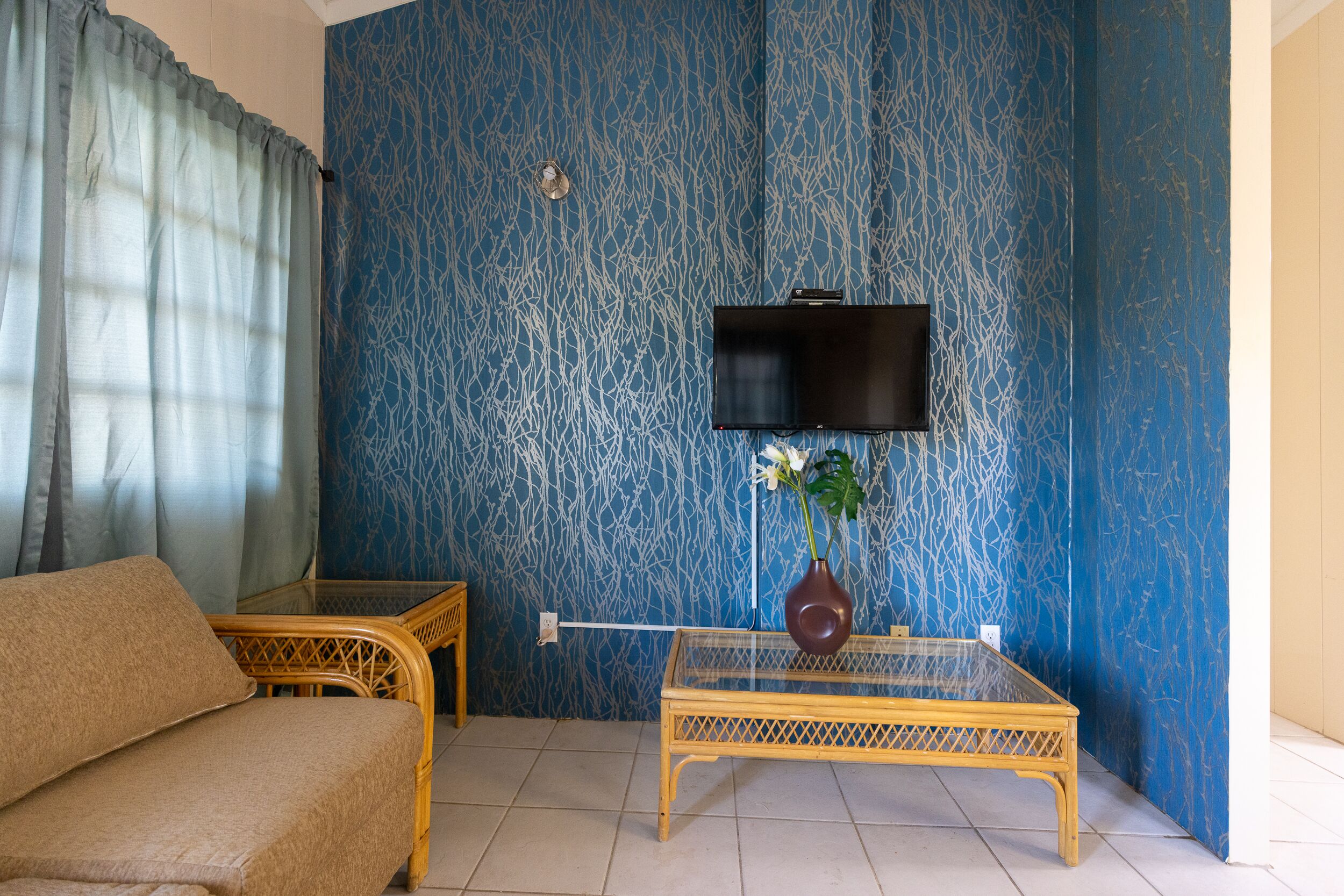 A room with a sofa, a glass-top table, a TV mounted on the wall, and a patterned blue wall.