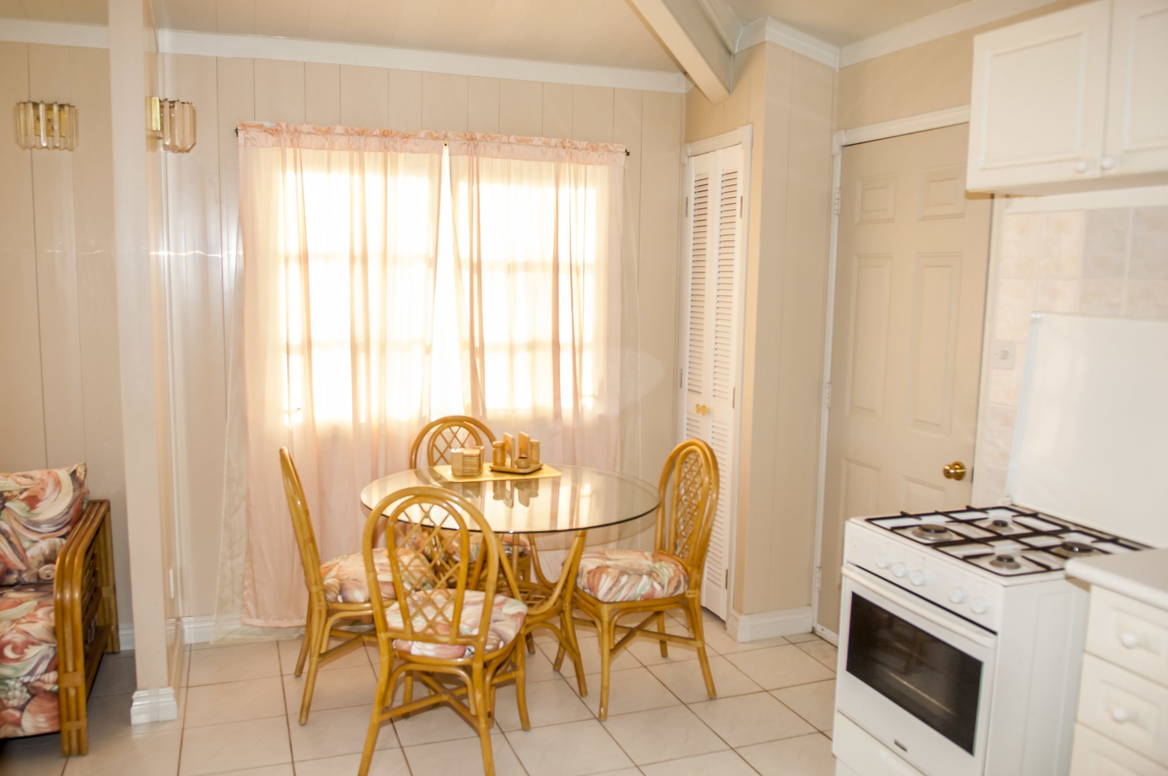 A dining area with a round glass table and four chairs, a window with sheer curtains, a white stove, and a door with louvered shutters.