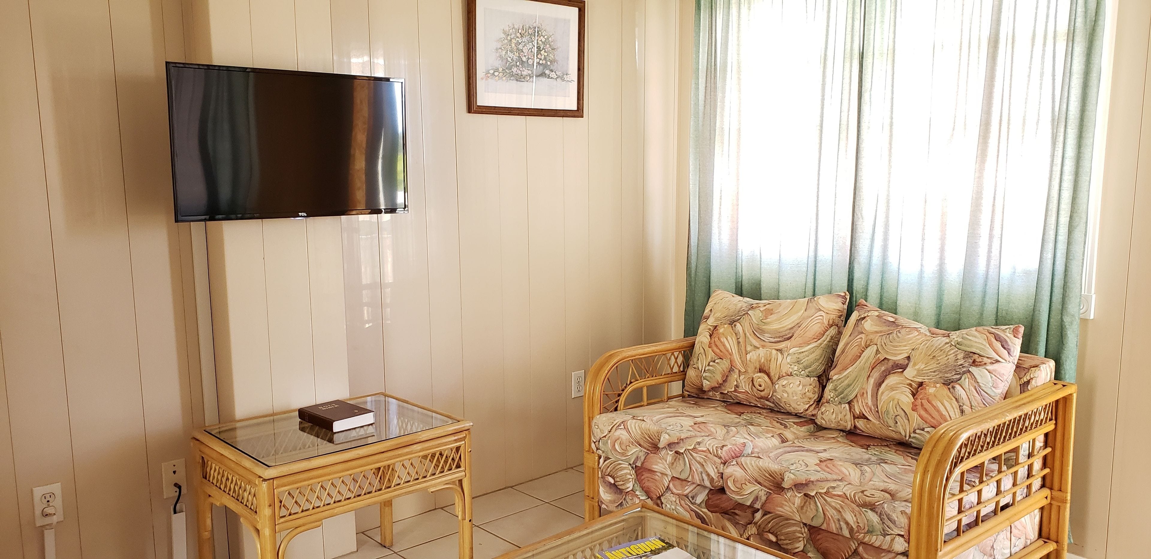 Un salon avec un téléviseur à écran plat fixé au mur, une table en verre sur laquelle repose un livre, un canapé à motifs et une fenêtre avec des rideaux.