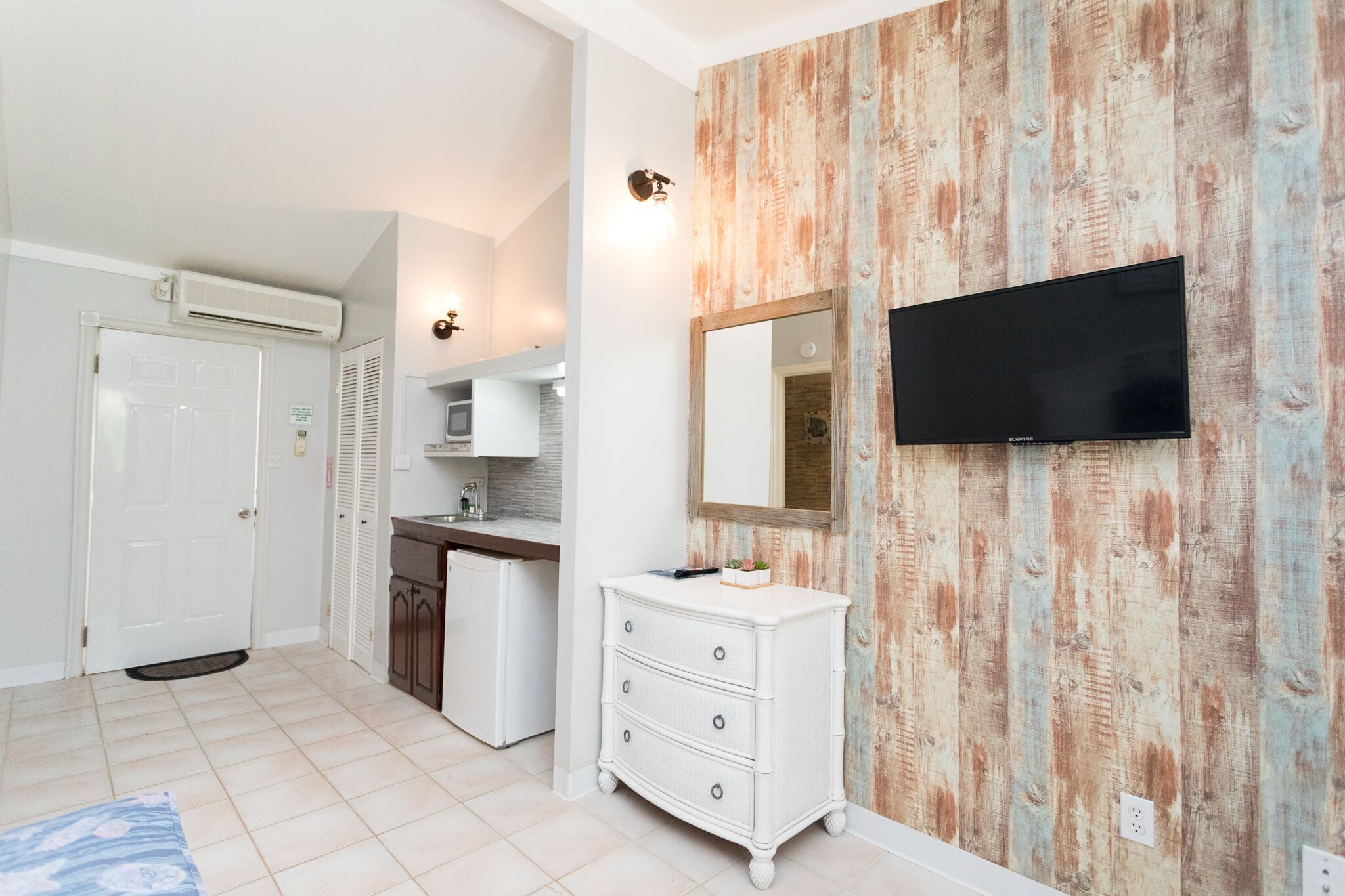 A hotel room with a wooden wall, a flat-screen TV, a white dresser, a door, and a kitchenette.