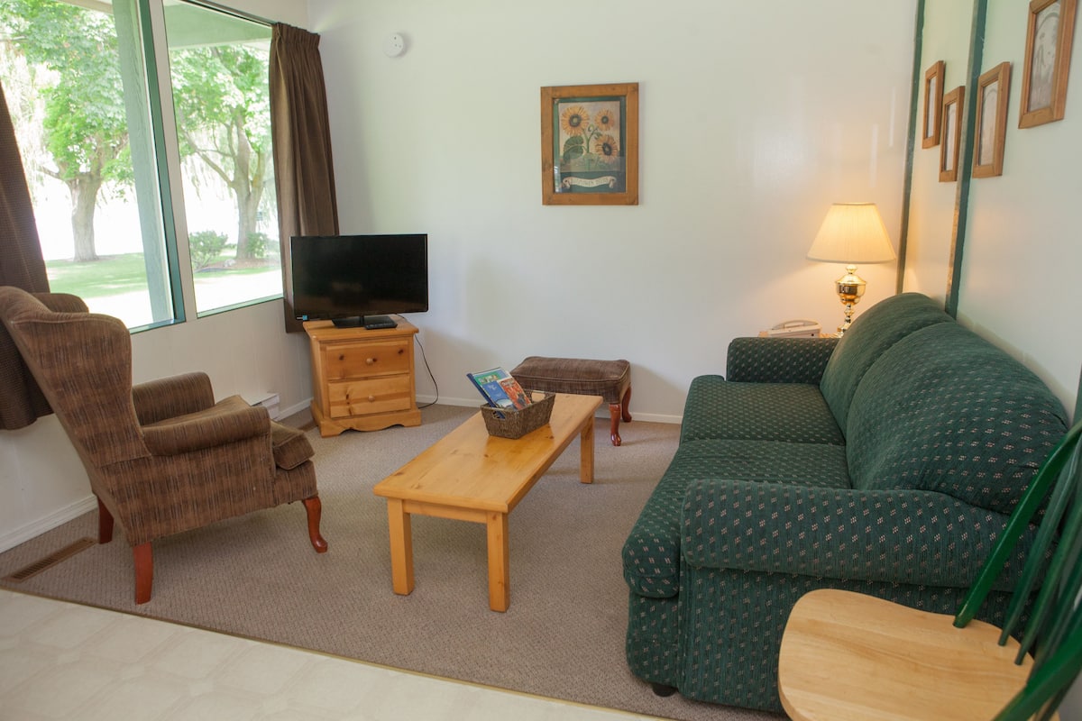 Una sala de estar con televisor, sofá, sillón, mesa de madera y lámpara.