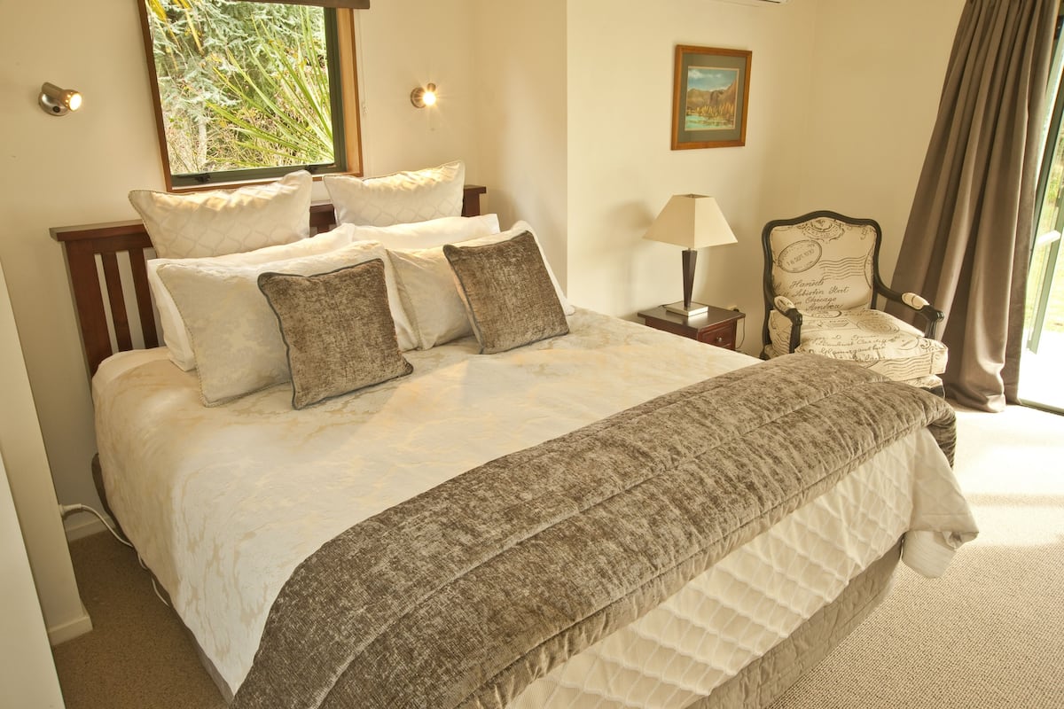 Uma cama bem arrumada com cobertura acolchoada e várias almofadas, uma mesa de cabeceira com abajur, uma cadeira com encosto decorativo e um quadro na parede.