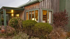 Une maison en bois à l’allure rustique, dotée d’un porche couvert et d’une porte vitrée donnant sur une pièce éclairée par une lampe.