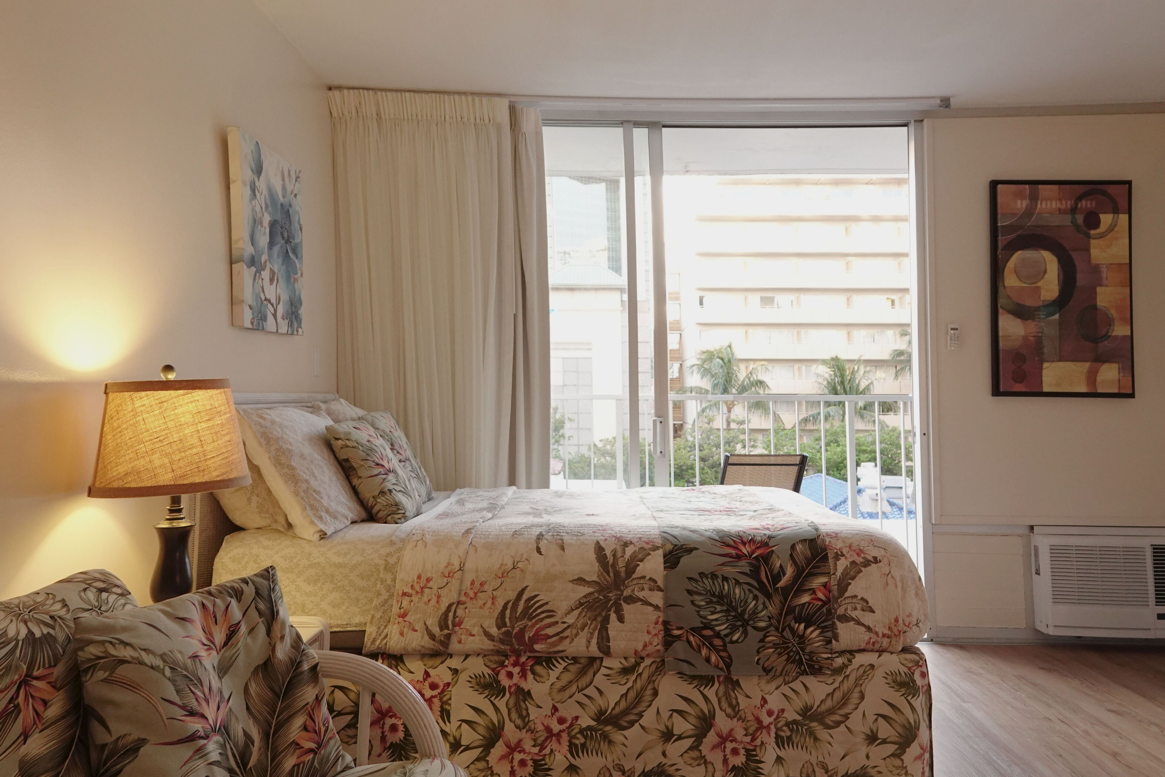 A hotel room with a bed, a nightstand, a lamp, a chair, a window with curtains, and a view of buildings and palm trees outside.