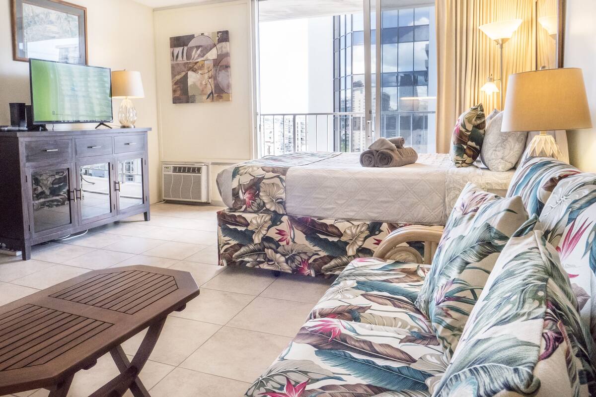 Hotellihuone, jossa on sänky, sohva, kaapissa oleva televisio ja parveke, jolta on näkymä.