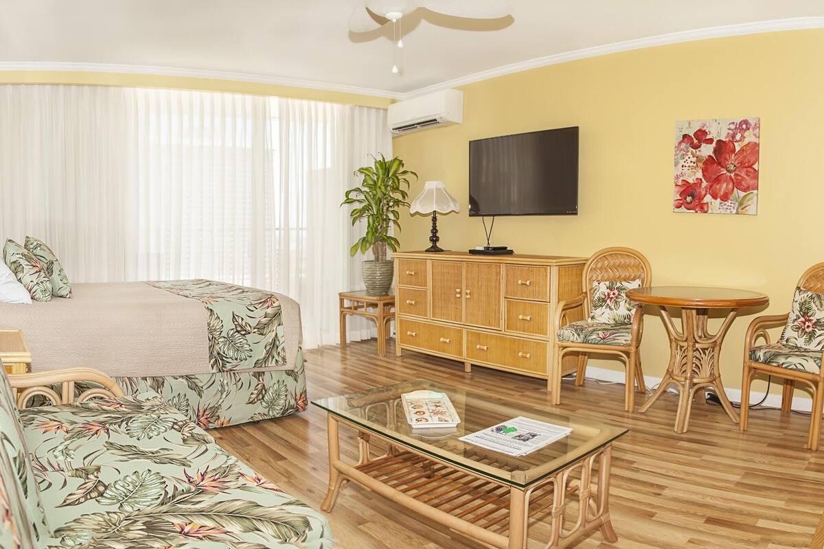 Hotellihuone, jossa on sänky, sohva, lasinen sohvapöytä, televisio, kaappi ja kaksi rottinkista tuolia.