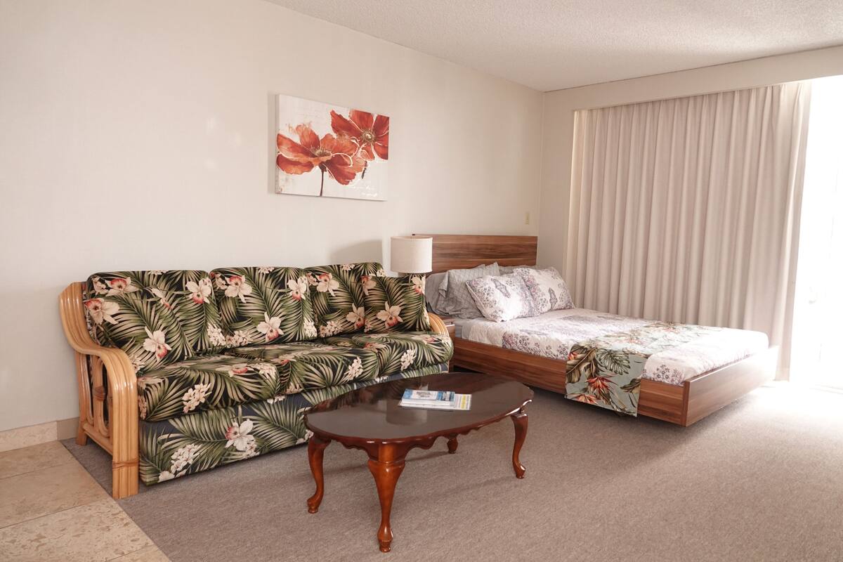 Hotellihuone, jossa on kukkakuviollinen sohva, puinen sohvapöytä, kukkakuviollinen päiväpeite sängyllä ja suuri ikkuna, jossa on verhot.