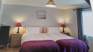 A double bed with two purple bedspreads, a nightstand with a lamp, a wall-mounted picture, and a window with curtains.