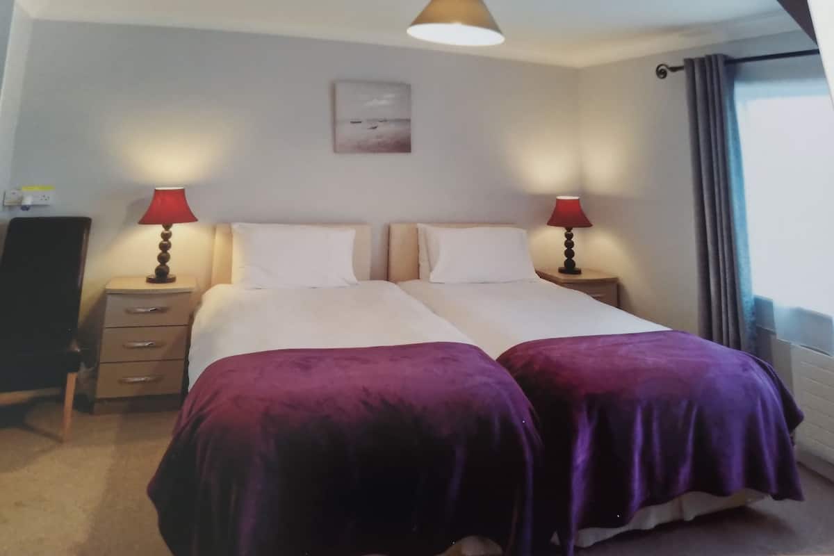 Un letto matrimoniale con due coperte viola, un comodino con una lampada, un quadro appeso al muro e una finestra con tende.