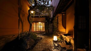 Un estrecho sendero de piedra que conduce a una puerta japonesa tradicional, rodeado de follaje y una lámpara pequeña.