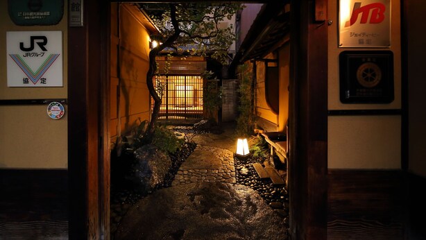 Un estrecho sendero de piedra que conduce a una puerta tradicional japonesa, flanqueado por muros de madera e iluminado por una cálida luz.