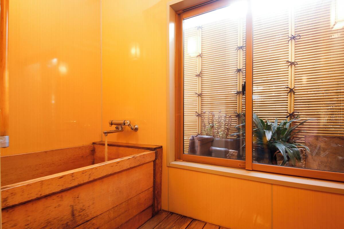 Un baño con una bañera de madera, una ventana con persianas de bambú y vistas a muebles de exterior.