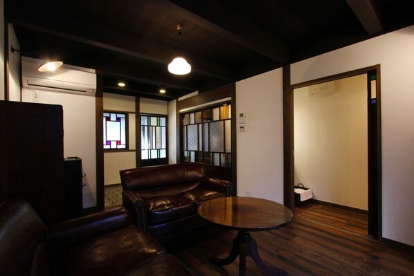 Quarto com sofá de couro, mesa redonda e piso de madeira. Há uma janela com vista para o exterior.