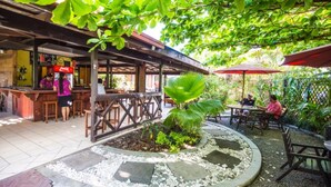 An outdoor cafe area with a wooden deck, tables, and chairs, surrounded by greenery and a decorative stone border. - Eden's Edge Hotel (Apia)