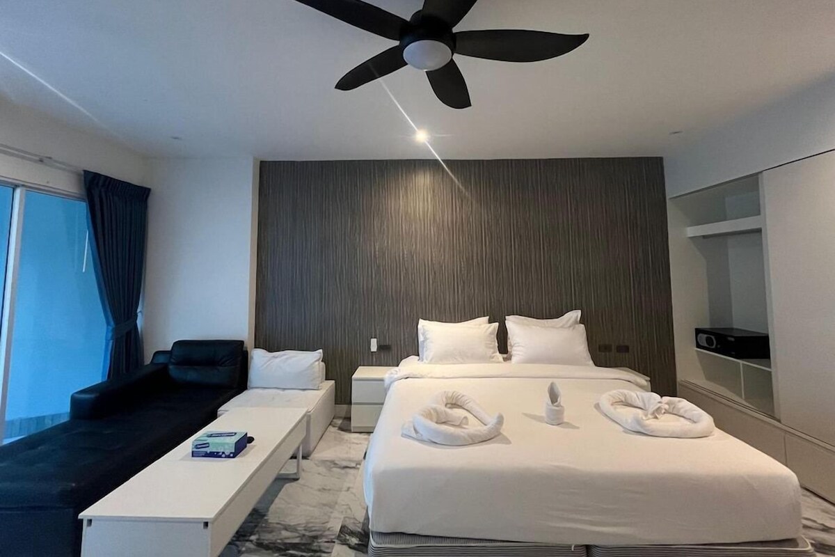 A hotel room with a bed, bedside tables, a desk, a chair, a ceiling fan, and a large window with curtains.