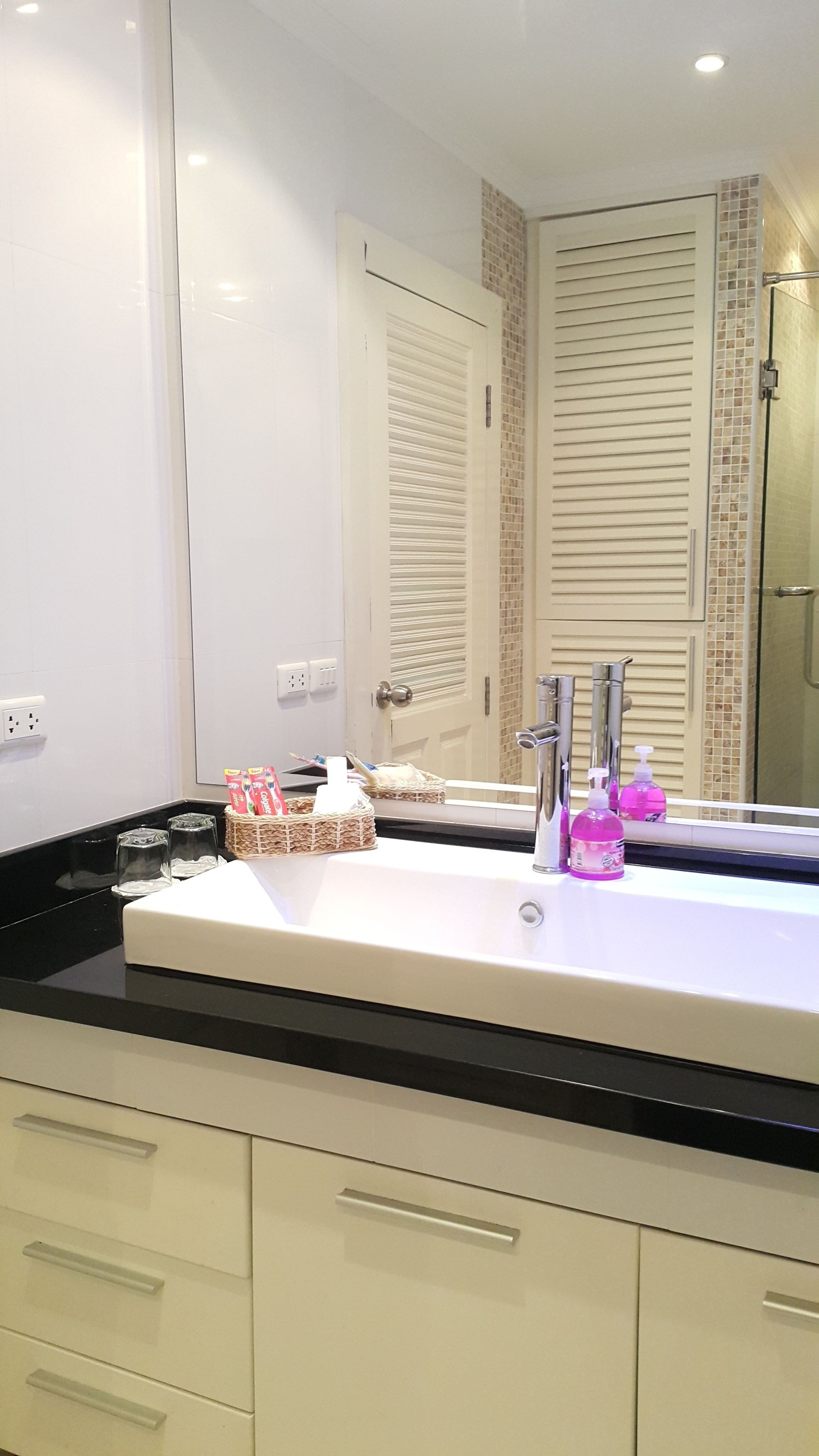 A bathroom with a large mirror, a white sink, and a black countertop. There are two bottles of hand soap and a basket with toiletries.