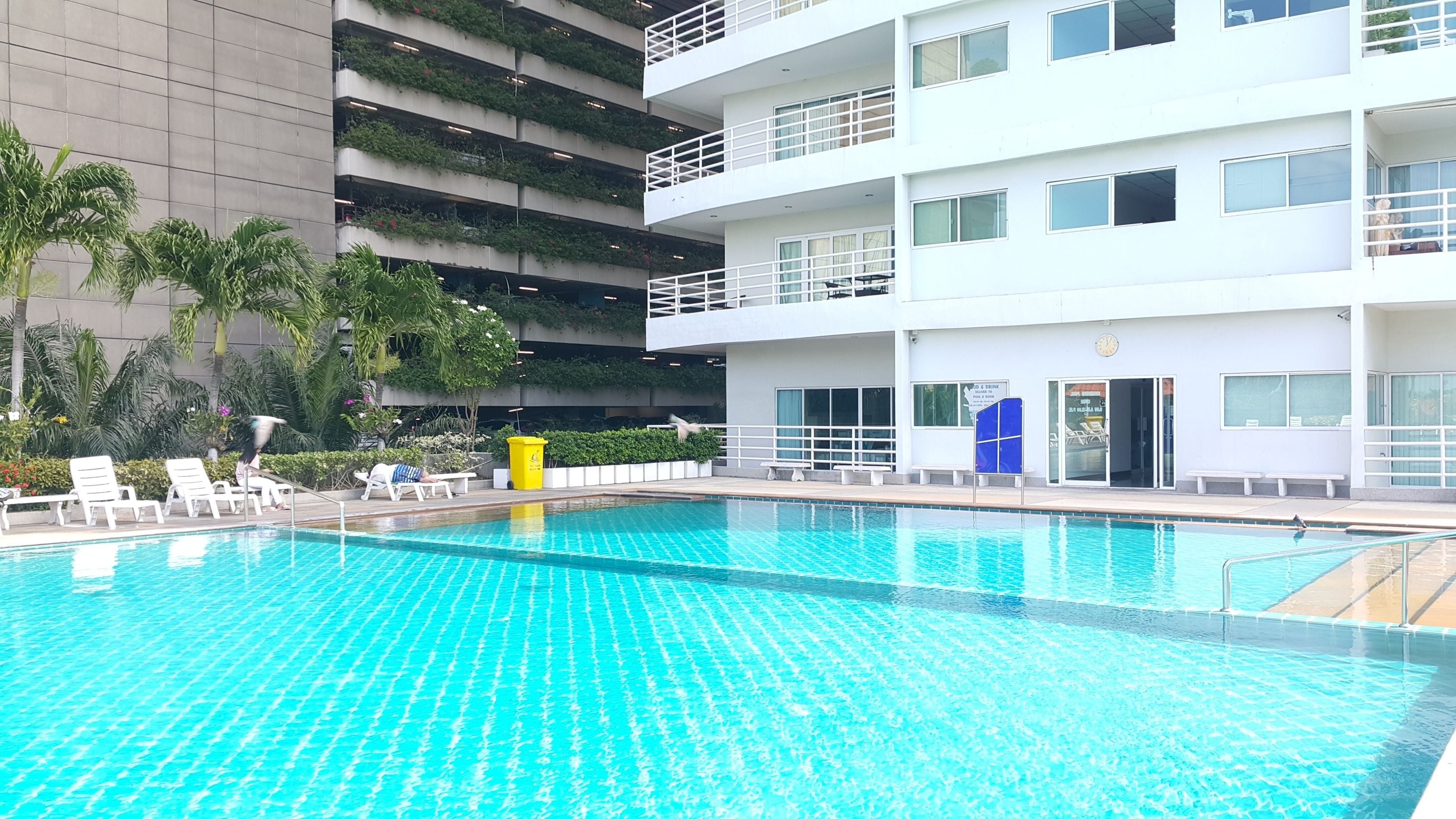A modern hotel with a swimming pool, multiple balconies, and lush greenery.