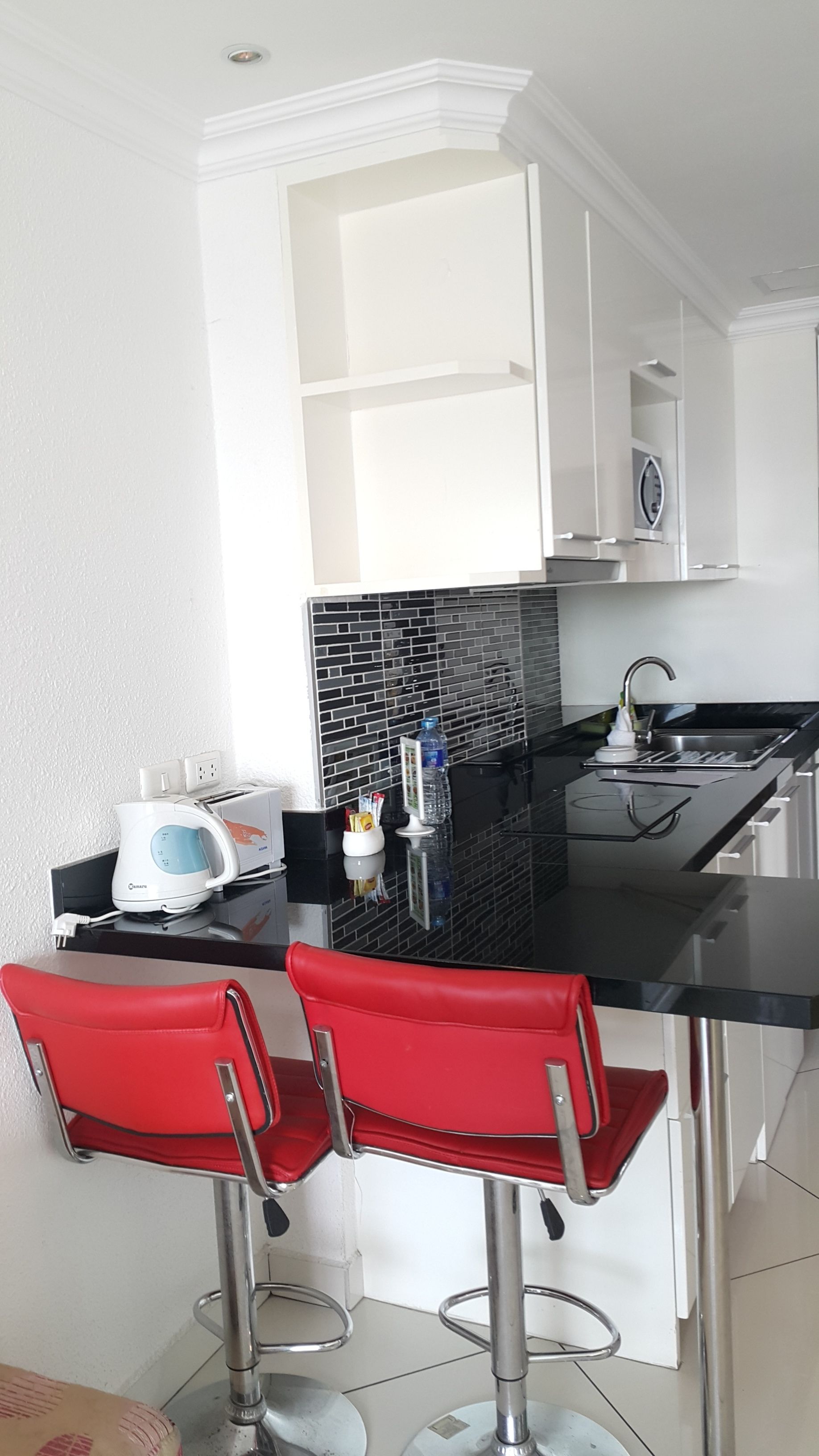 Een moderne keuken met witte kasten, een zwarte achterwand en een roestvrijstalen spoelbak. Op het aanrecht staan twee rode barkrukken en een witte waterkoker.