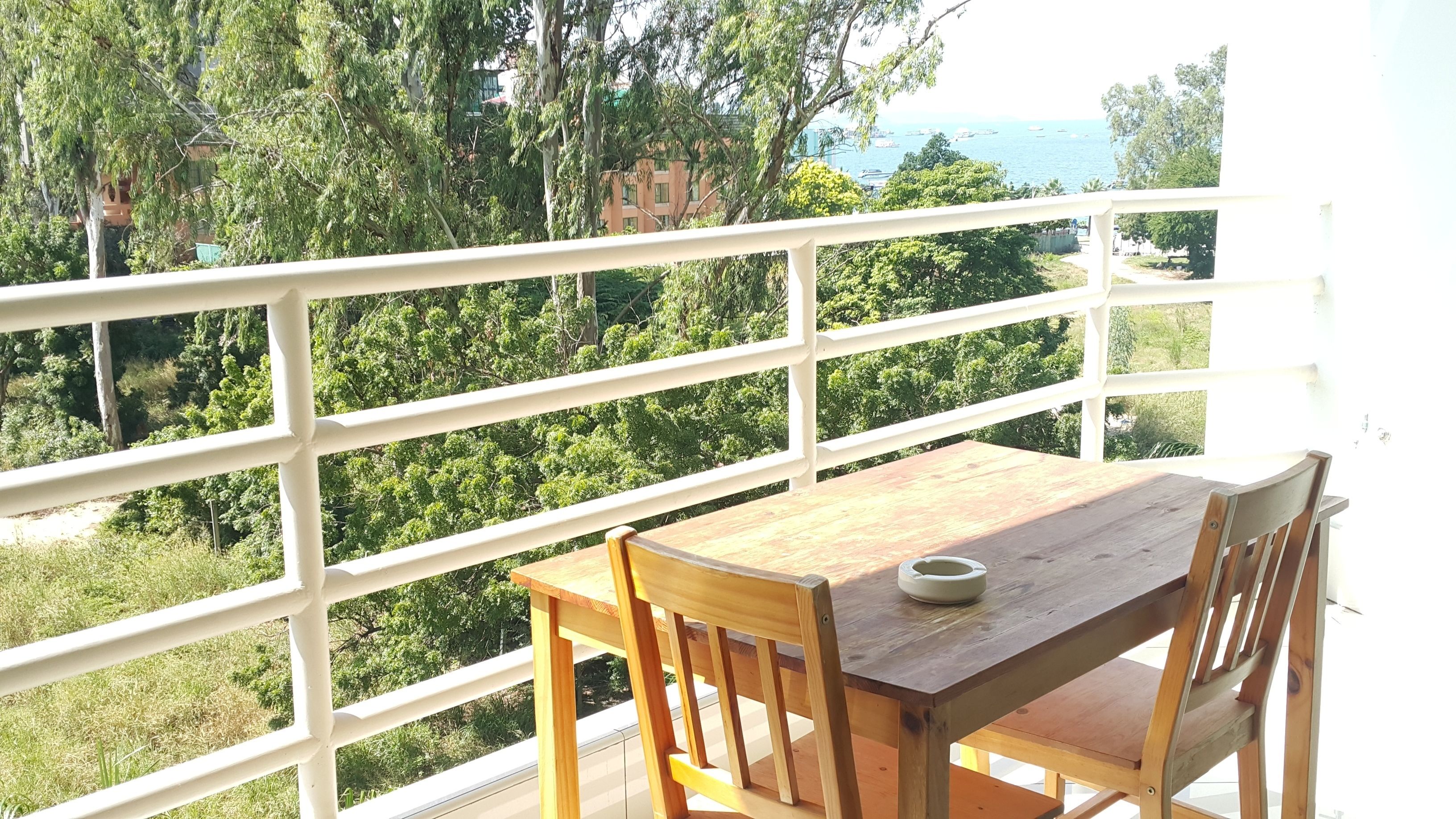 A wooden table and chair on a balcony with a white railing, overlooking a lush green landscape and a body of water with boats.