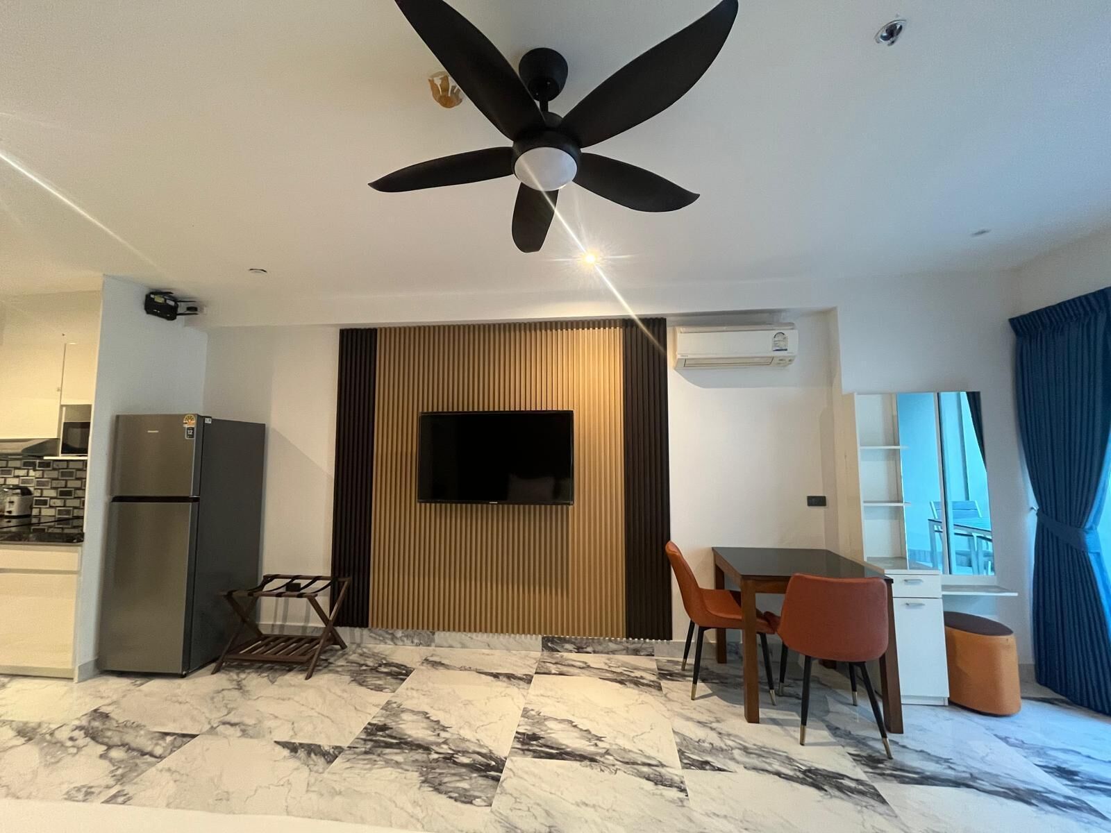 A modern living room with a ceiling fan, a flat-screen TV, a dining area with chairs, a kitchen with a refrigerator, and a patterned floor.