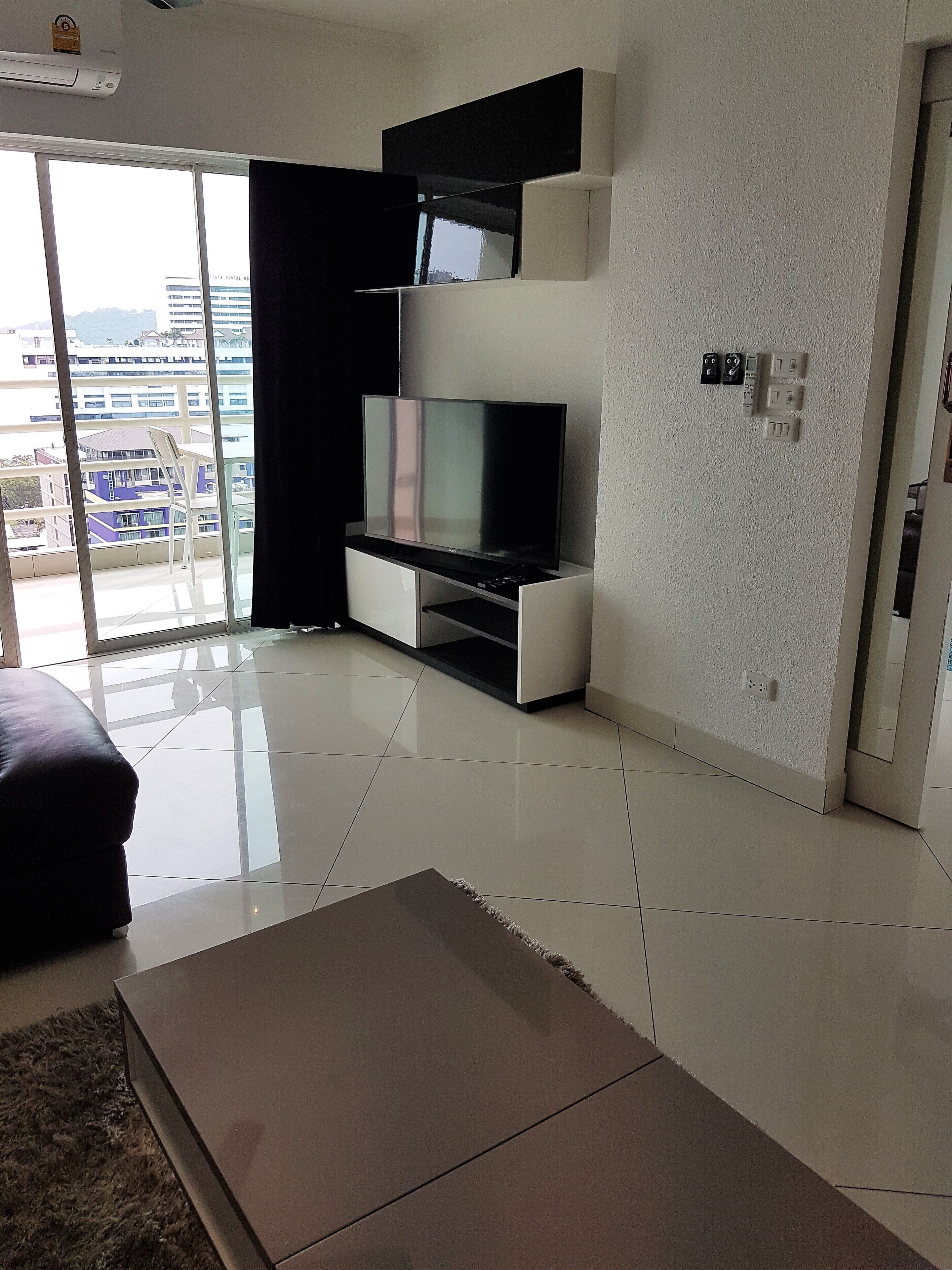 Ein modernes Wohnzimmer mit einem Flachbildfernseher auf einem weißen Schrank, einer schwarzen Couch und einem großen Fenster mit Blick auf Gebäude und Berge.