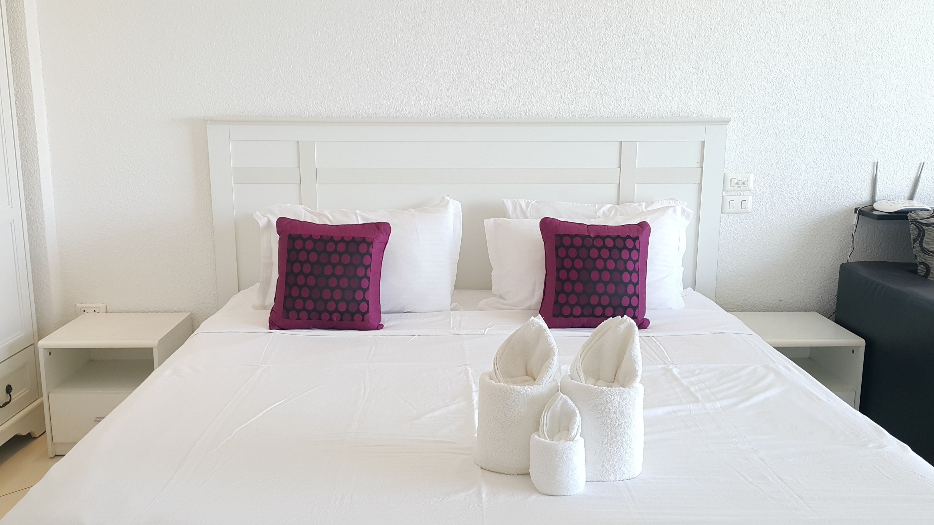 Una cama bien hecha con ropa blanca y dos almohadas moradas con estampado de lunares. Hay dos pantuflas blancas sobre la cama.