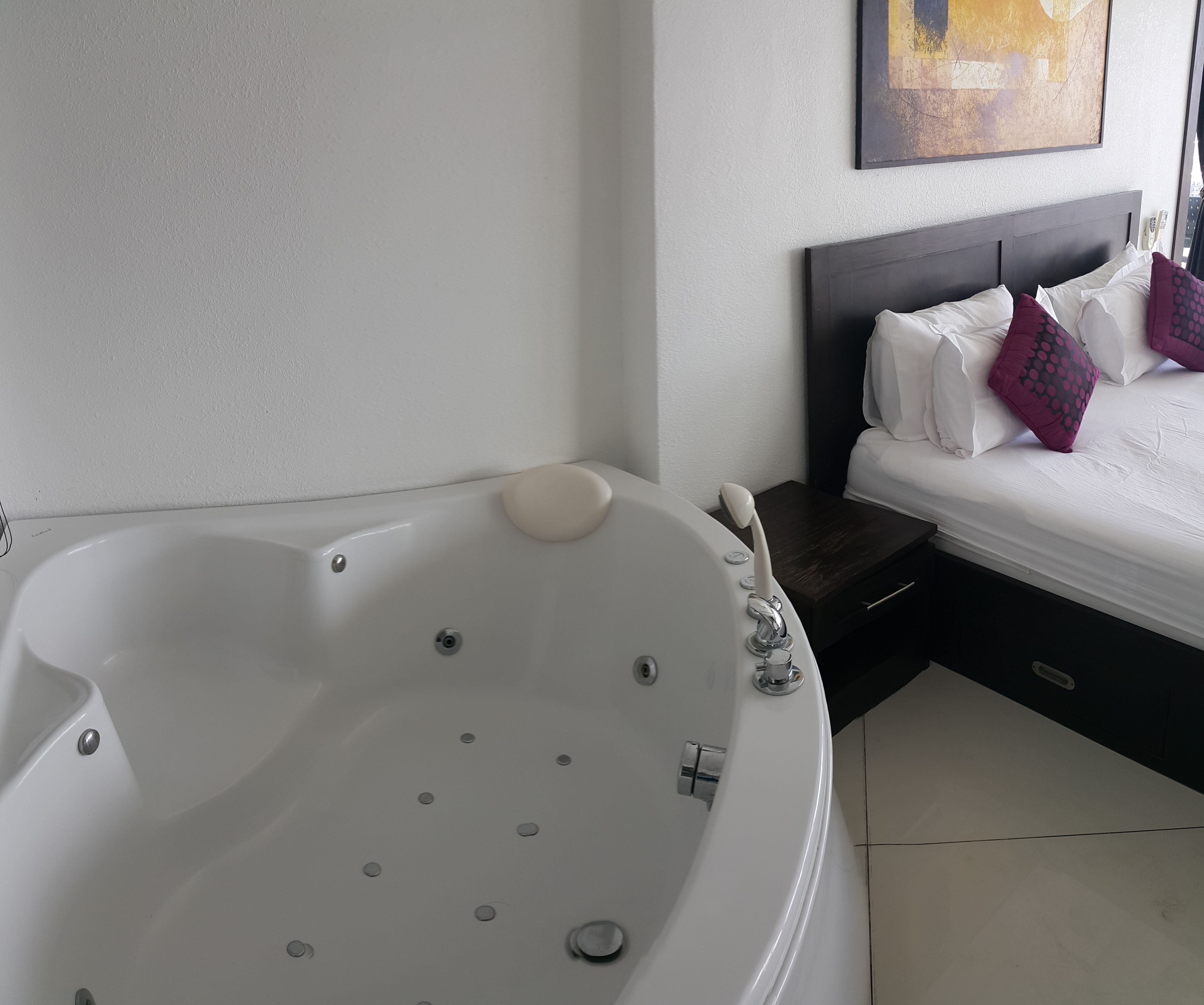 A modern bathroom with a freestanding bathtub, a bedside table, and a bed with white bedding and decorative pillows.