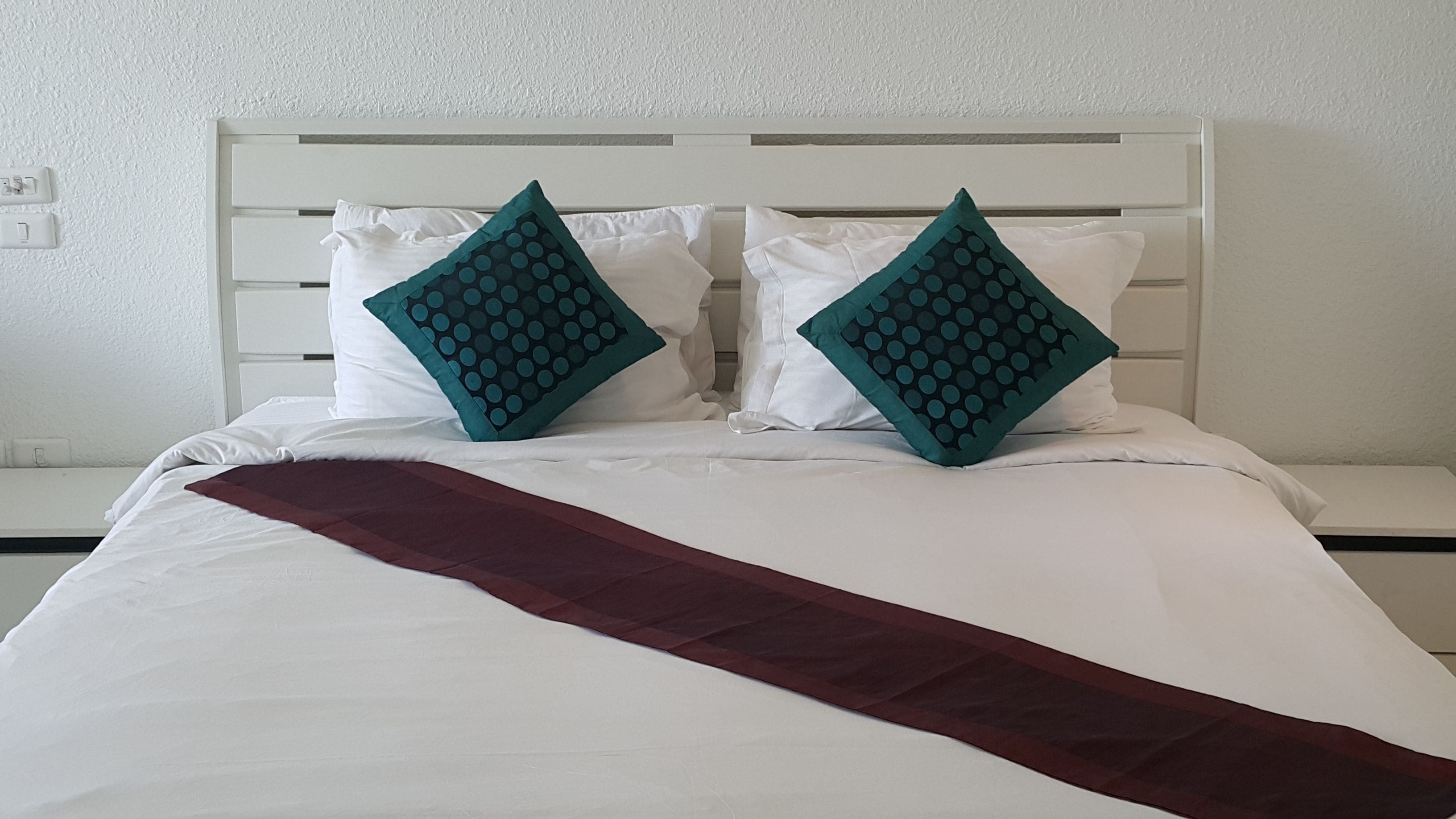 Un lit avec une literie blanche, deux coussins turquoise à motifs à pois et un plaid bordeaux.