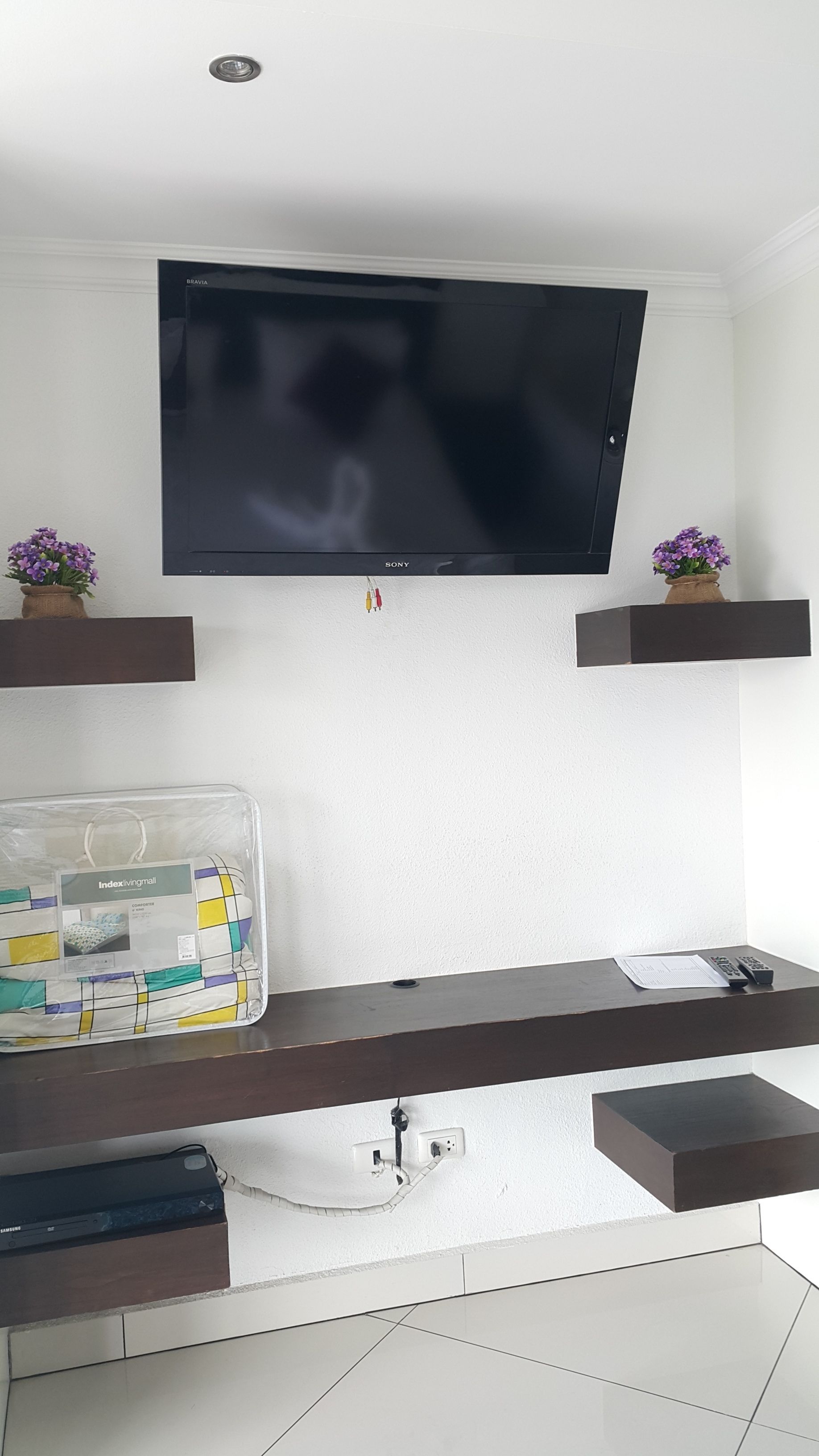 Una sala de estar moderna con un televisor de pantalla plana montado en la pared, un mueble de madera debajo y dos pequeños arreglos florales en estantes flotantes.