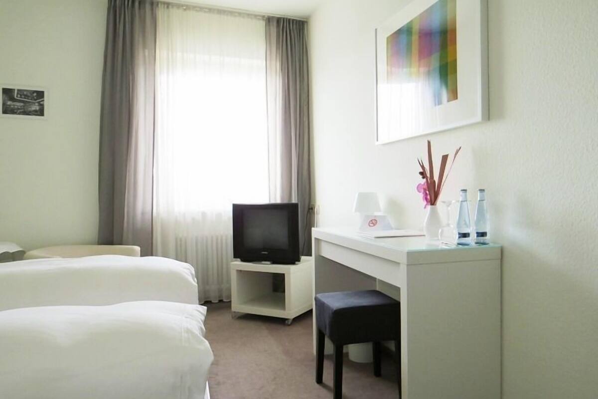 A hotel room with two beds, a desk with a TV, a small table with a lamp and decorative items, and a window with curtains.
