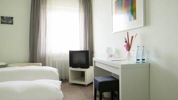 Habitación de hotel con dos camas, un escritorio con televisor, una mesita con lámpara y objetos decorativos, y una ventana con cortinas.