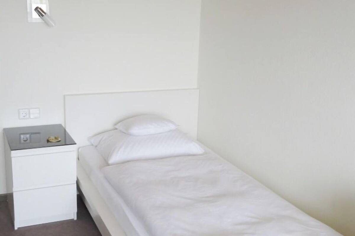 A small, single-bed room with a white bed, a nightstand, and a wall socket.