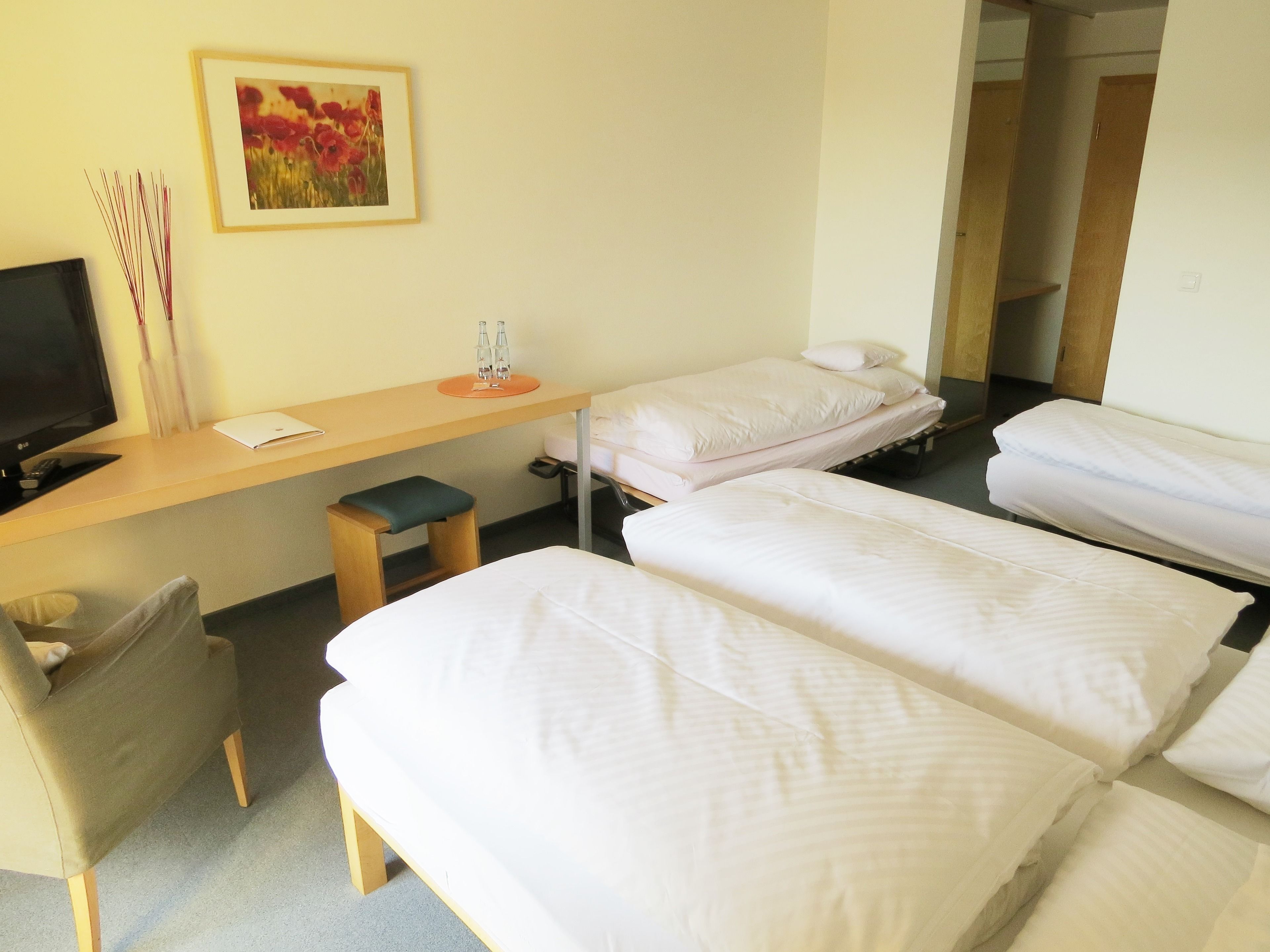 A hotel room with two single beds, a desk with a computer, a chair, and a small table with a vase and bottles.