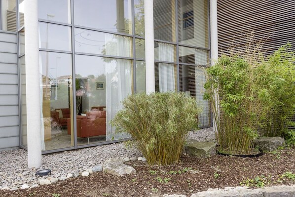 Un edificio moderno con grandes ventanales, un jardín con arbustos y rocas, y un interior visible con mobiliario.