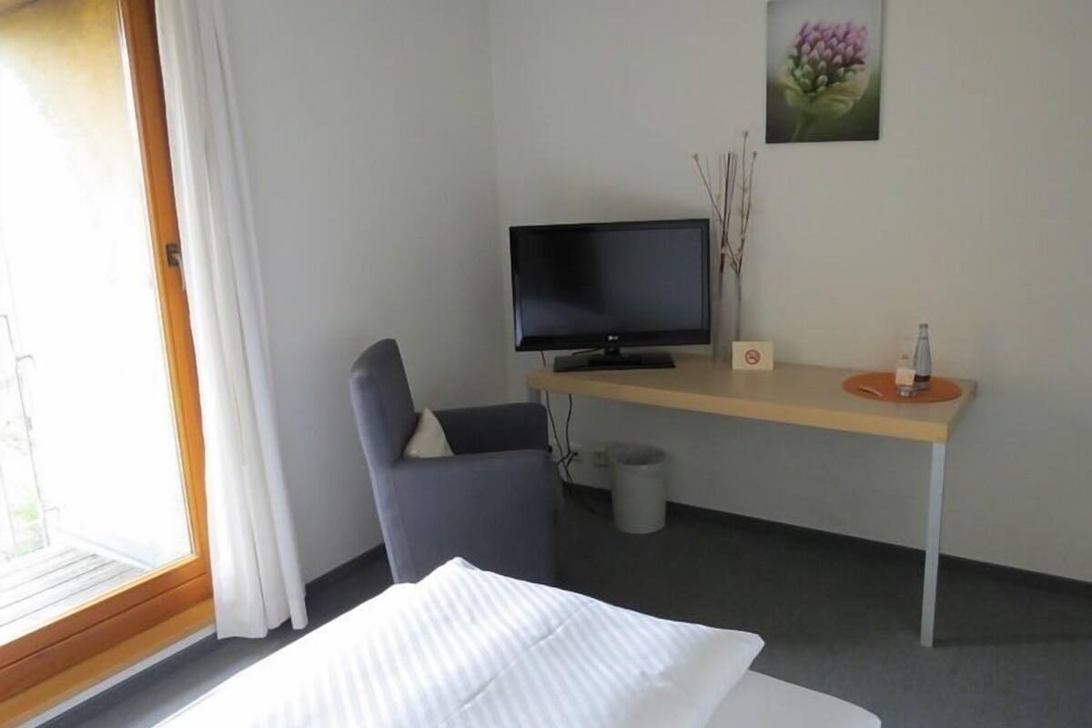 A hotel room with a bed, a desk with a television, a chair, a small table with a bottle, and a picture on the wall.