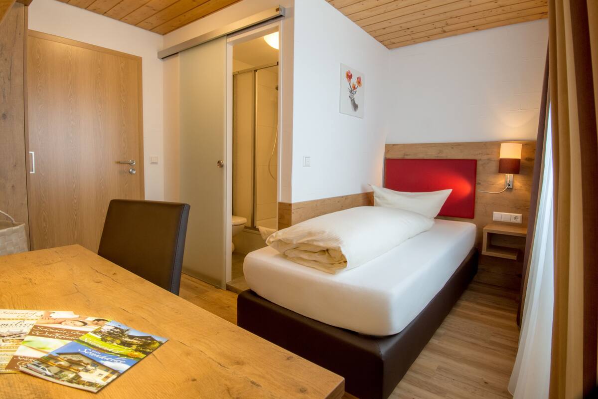 A bedroom with a wooden floor, a bed with white bedding, a wooden nightstand with a lamp, a wooden door, and a wall-mounted picture.