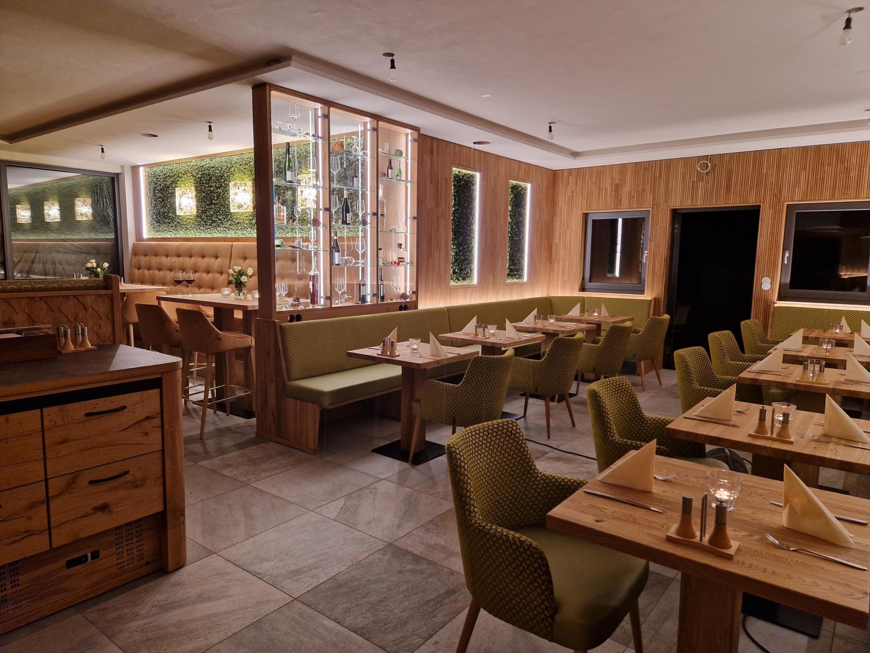 Un restaurante con mobiliario de madera, asientos acolchados verdes y una vitrina de vidrio con botellas de vino.