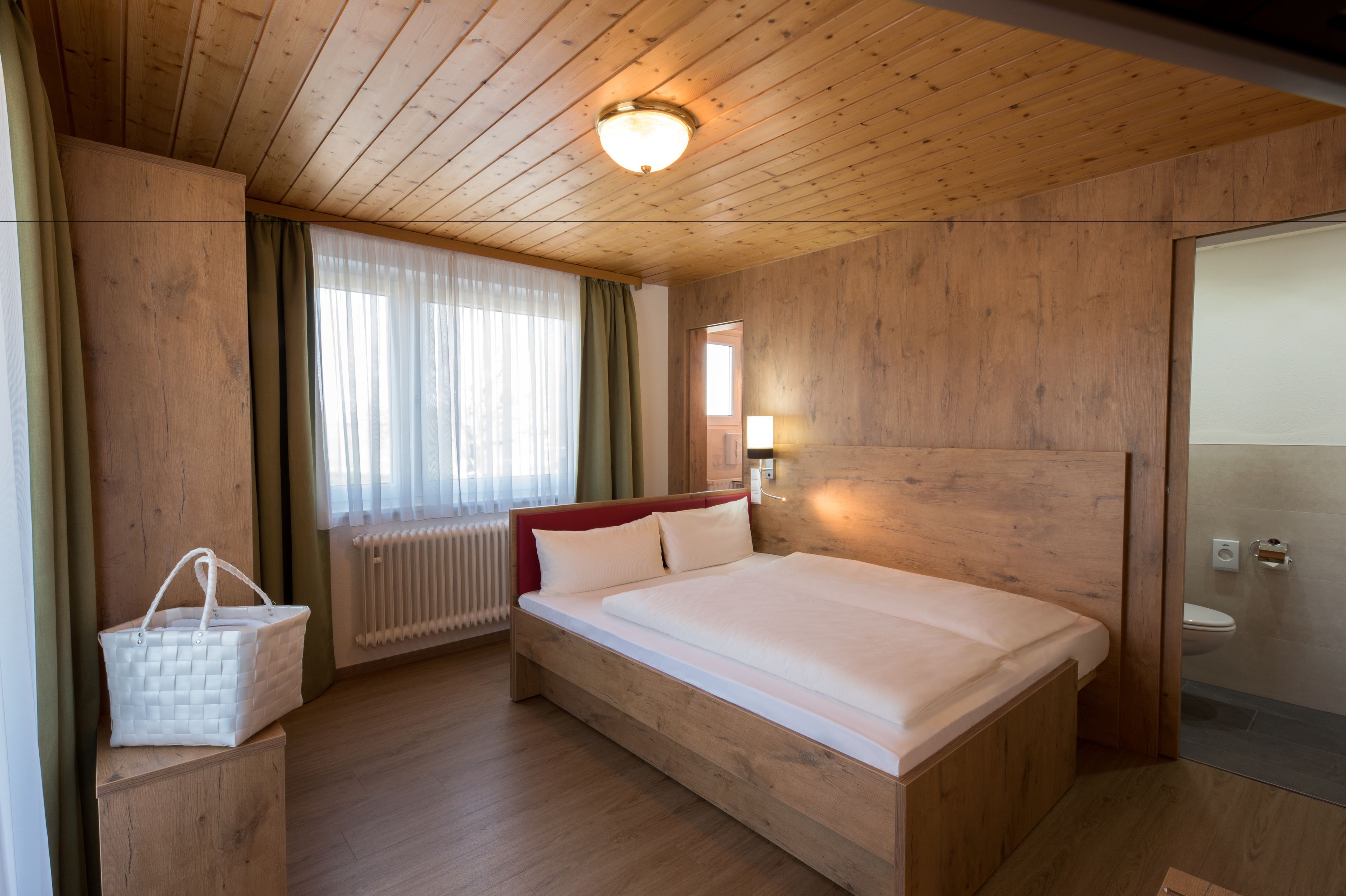 Une chambre à coucher aux murs lambrissés, avec un lit, un radiateur, une table de chevet, un sac à main blanc et une fenêtre avec des rideaux.