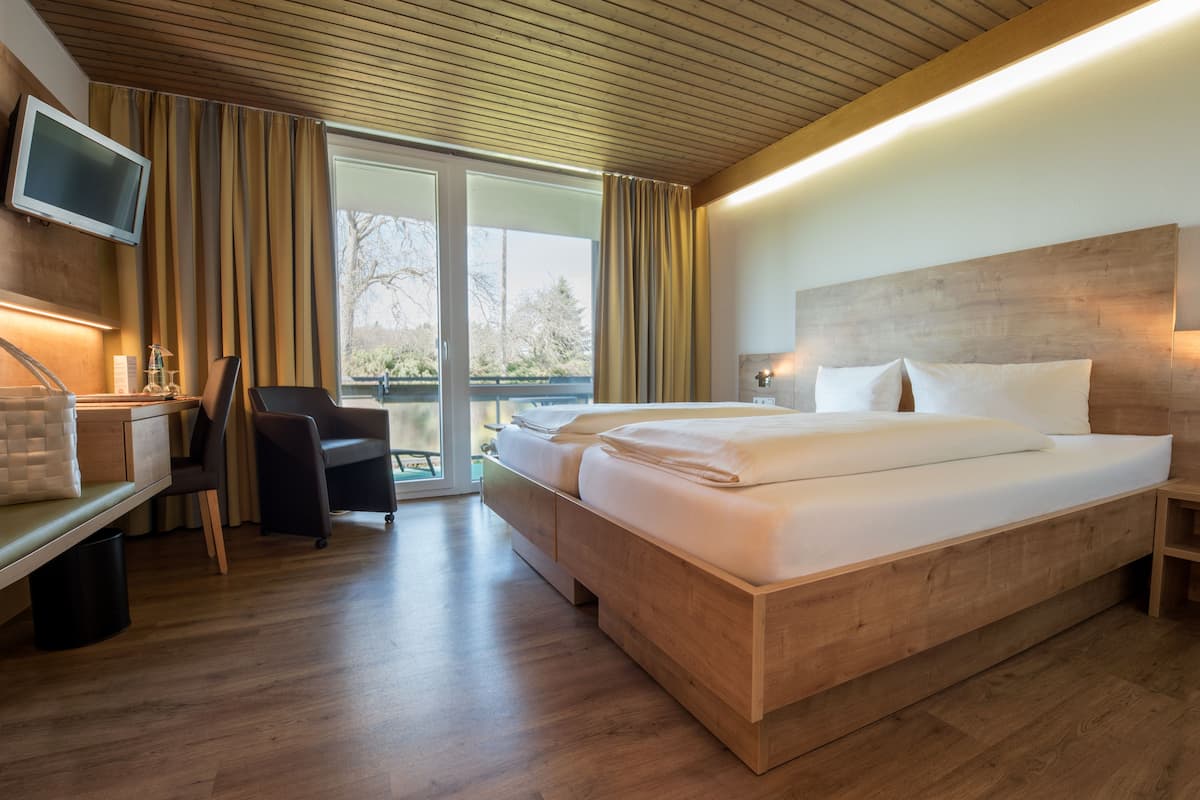 A modern hotel room with a wooden bed, a desk with a chair, a television, and a large window with curtains.