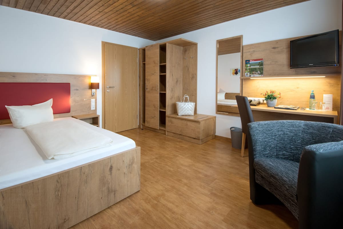 A modern hotel room with a wooden headboard, a bed, a desk with a chair, a wardrobe, and a television.
