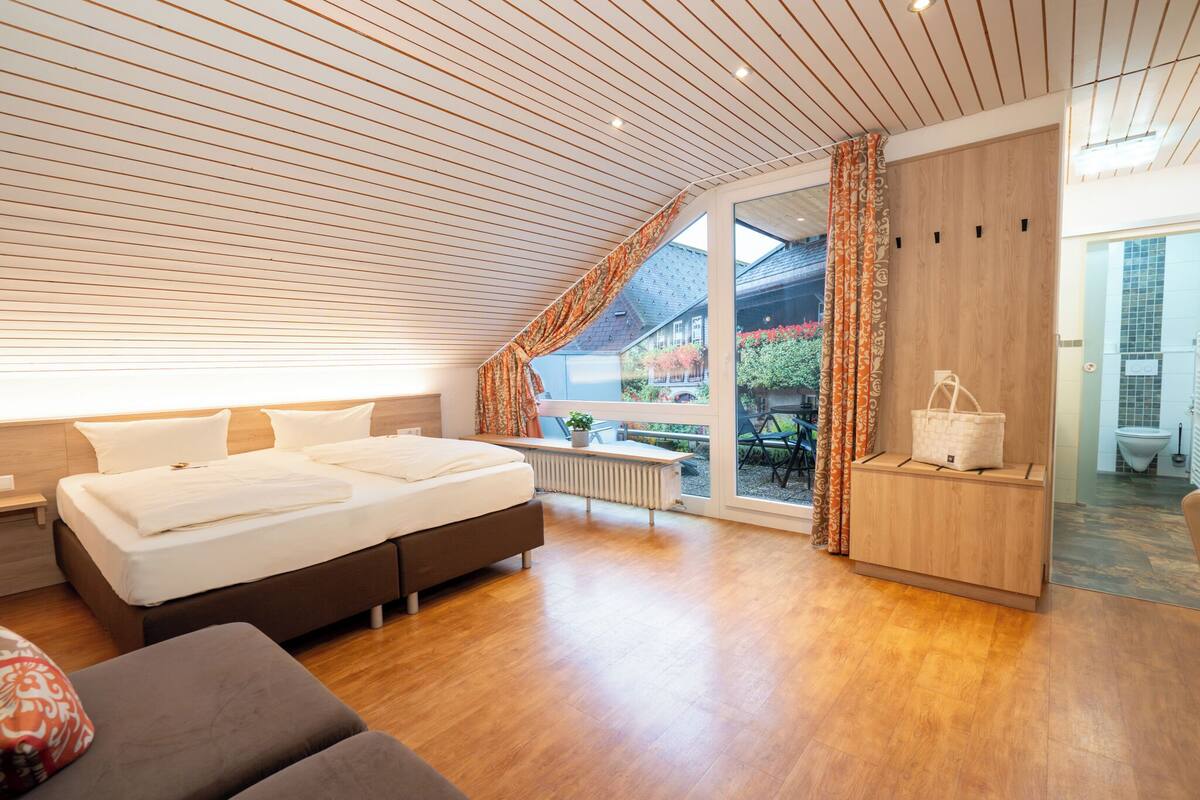 A modern bedroom with a large bed, wooden ceiling, and a view of the garden through a sliding door.