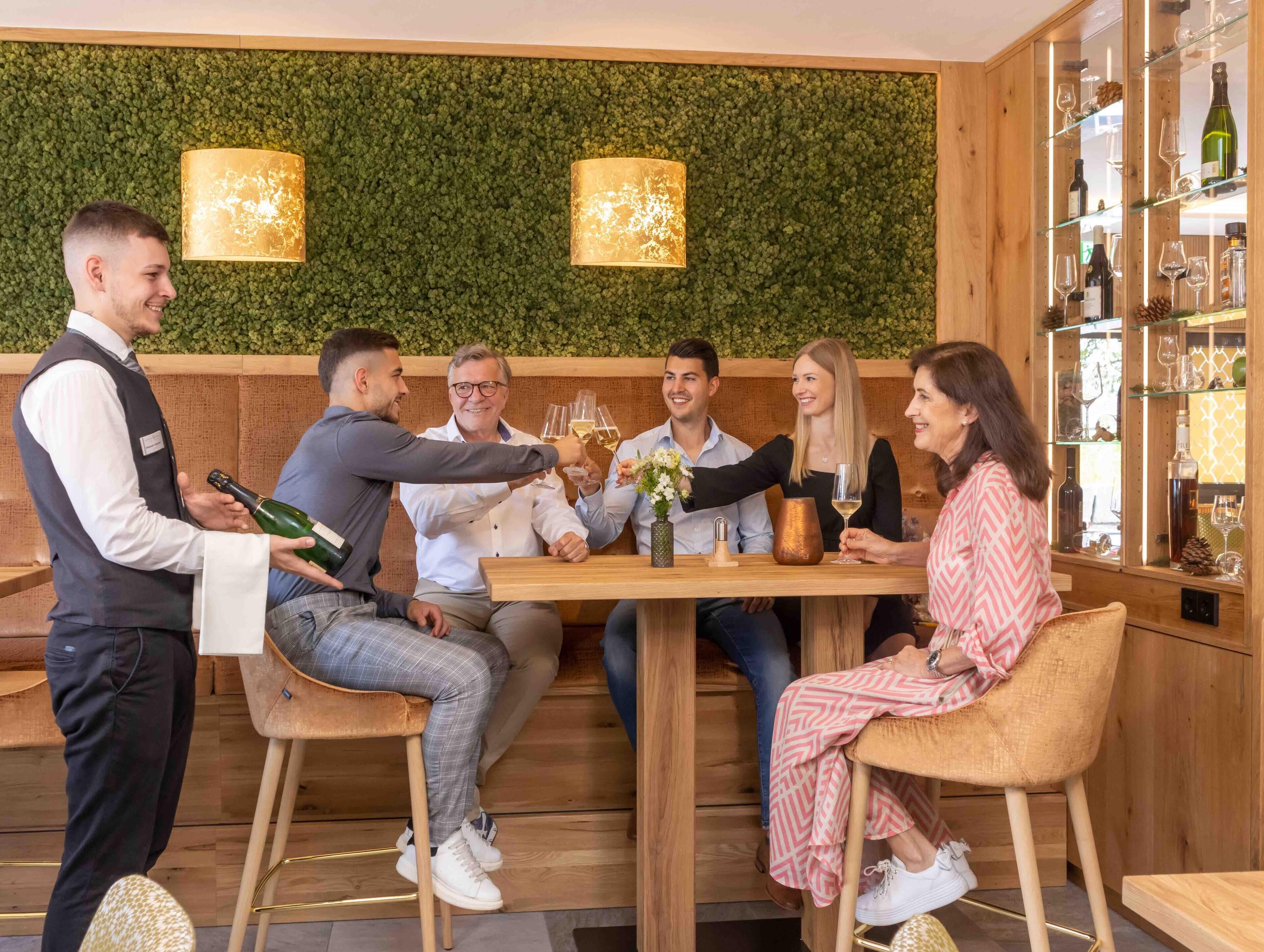 A group of people seated around a wooden table in a restaurant with a green wall and two hanging lamps.