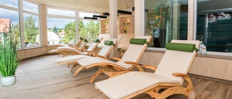 A sunlit room with wooden lounge chairs and large windows offering a view of the outdoors.