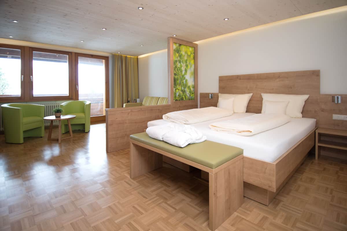 A modern hotel room with a wooden bed, a bench, a green chair, and a view of the outdoors through large windows.