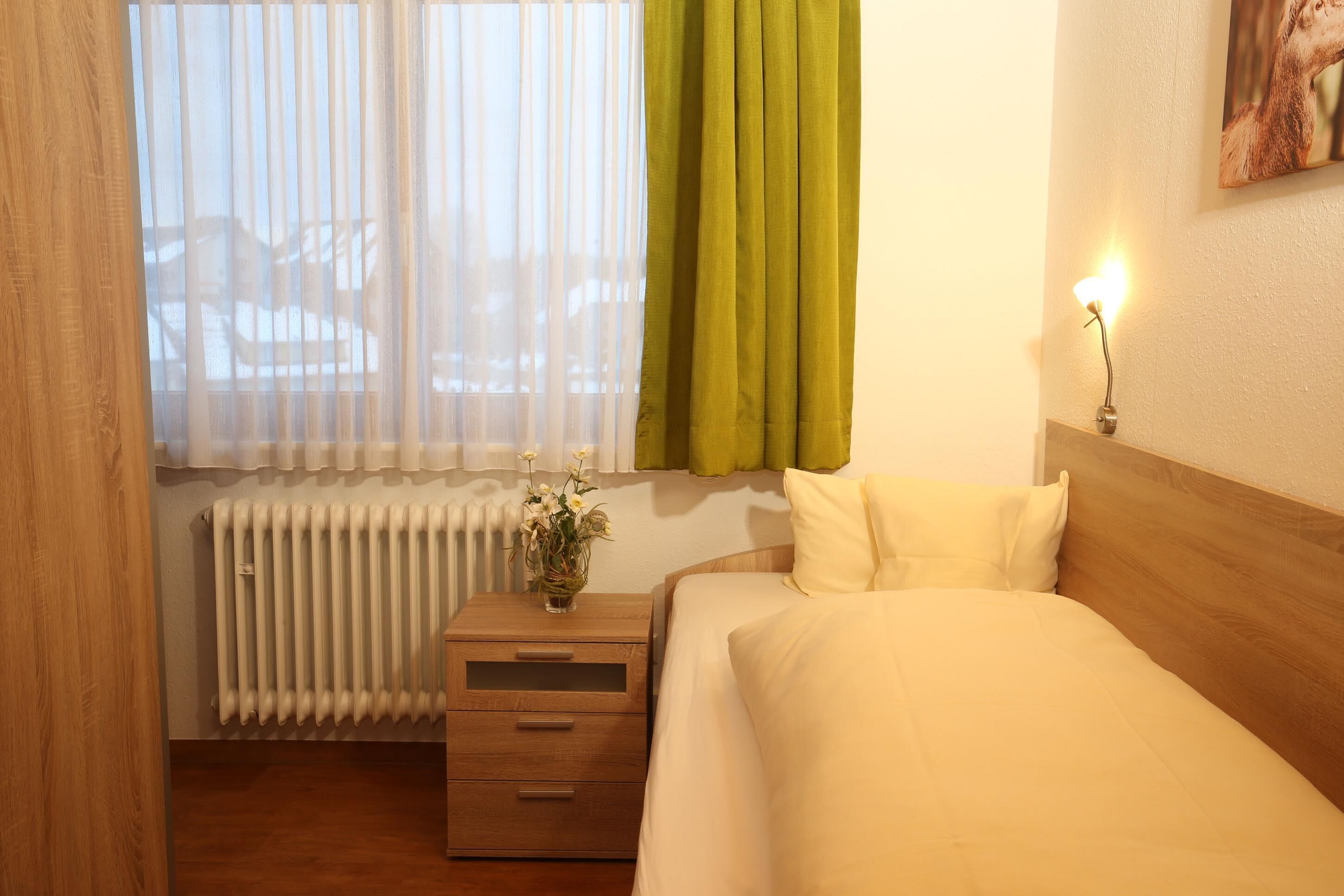 A hotel room with a bed, a nightstand with a vase of flowers, a radiator, and a window with sheer curtains and a green drape.