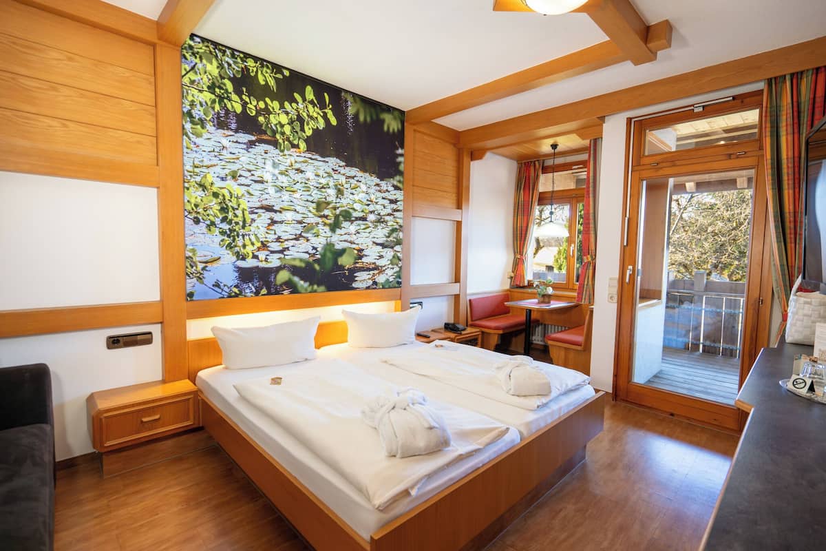 A hotel room with a large bed, wooden walls, a painting of a pond, a small table, and a balcony with curtains.